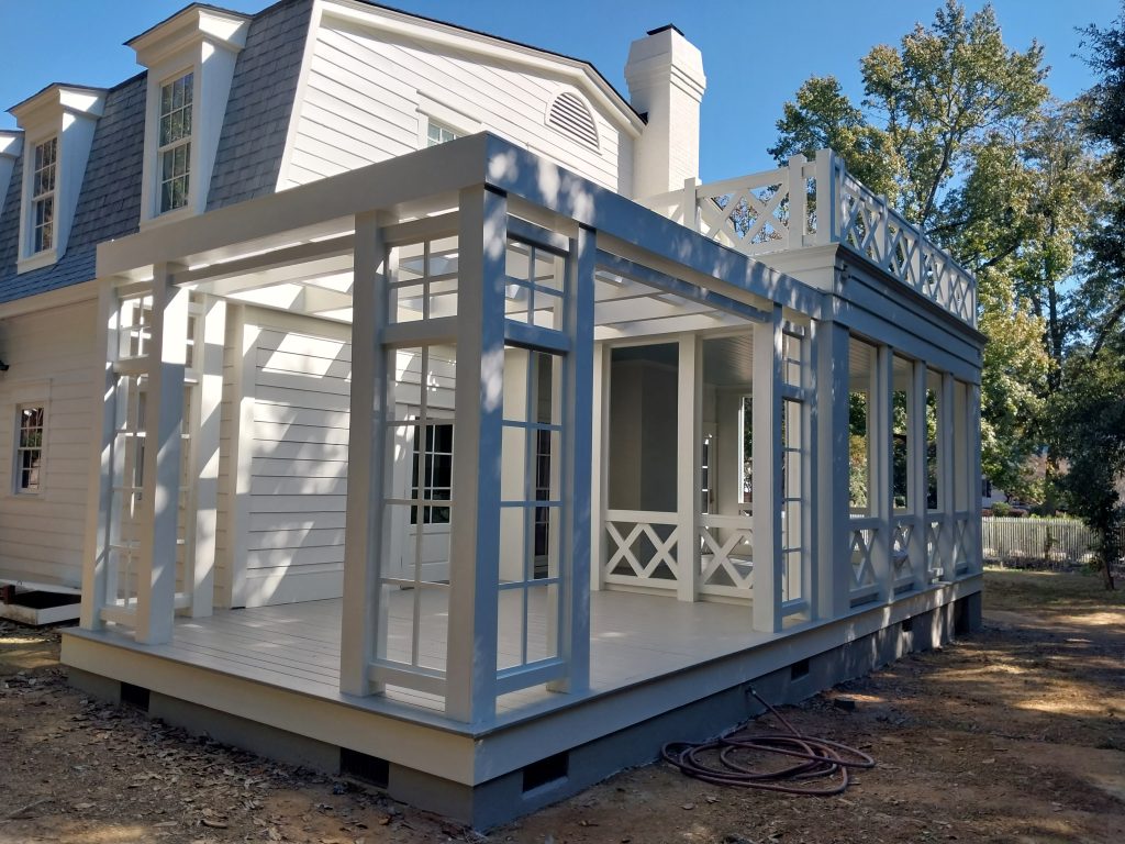 Residential sunroom addition under construction featuring white trim framing and blue-gray supports with glass doors alongside traditional home.